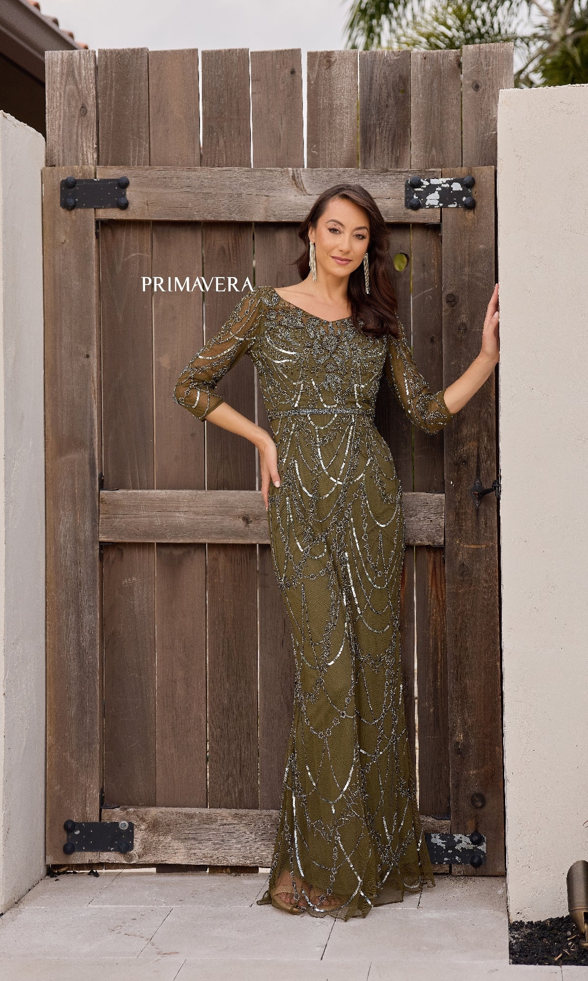 A woman in the Primavera 13156 long formal dress, featuring olive green sequins and a beaded waistline, stands before a rustic wooden gate labeled